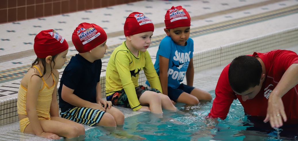 Children in red caps sit at the pool's edge during a swimming lesson.