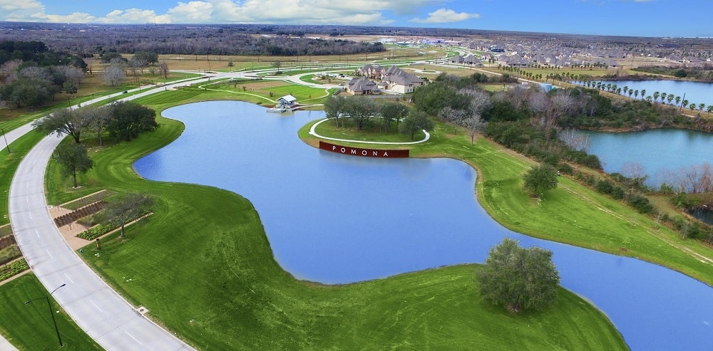 An aerial drone view of the Pomona master-planned community in Manvel, Texas, featuring a large bright blue amenity pond with a curved "POMONA" sign, lush green parks, winding roads, and luxury homes in the distance under a clear sky.