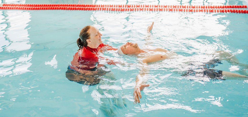 Smiling swim instructor gently supports a man floating on his back in a bright blue pool. TITLE