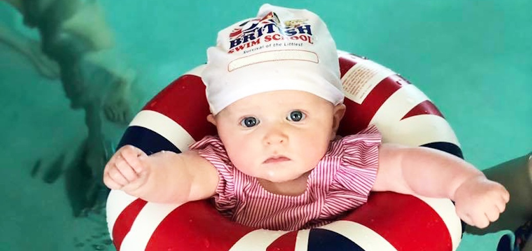 A baby wearing a British Swim School cap and a Union Jack float stares into the camera.