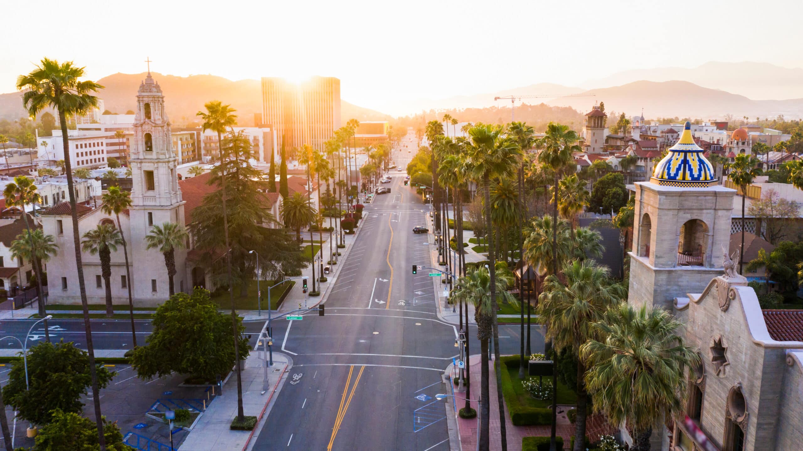 A panoramic aerial view of historic downtown Riverside, California, captured at sunrise, featuring the landmark Mission Inn Hotel & Spa with its ornate, eclectic architecture and the bell tower of the Riverside First United Methodist Church bordering a quiet, palm-lined street. British Swim School offers year-round, indoor swim lessons for the Riverside County community and surrounding Riverside, Coronado, Jurupa Valley, and Moreno Valley neighborhoods.