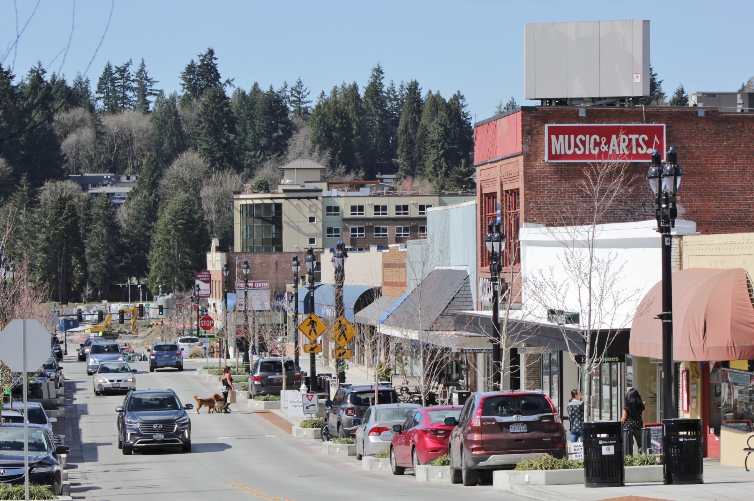 A street-level view of Main Street in Bothell, Washington, featuring a mix of historic brick and modern buildings under a clear sky. A "Music & Arts" sign is prominent on a red brick building to the right, while cars drive along the road and pedestrians walk dogs on the sidewalk. Dense evergreen trees rise on a hill in the background.