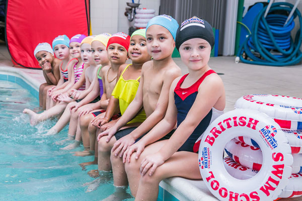 A group of smiling children in swim caps and suits sit by a pool with British Swim School rings