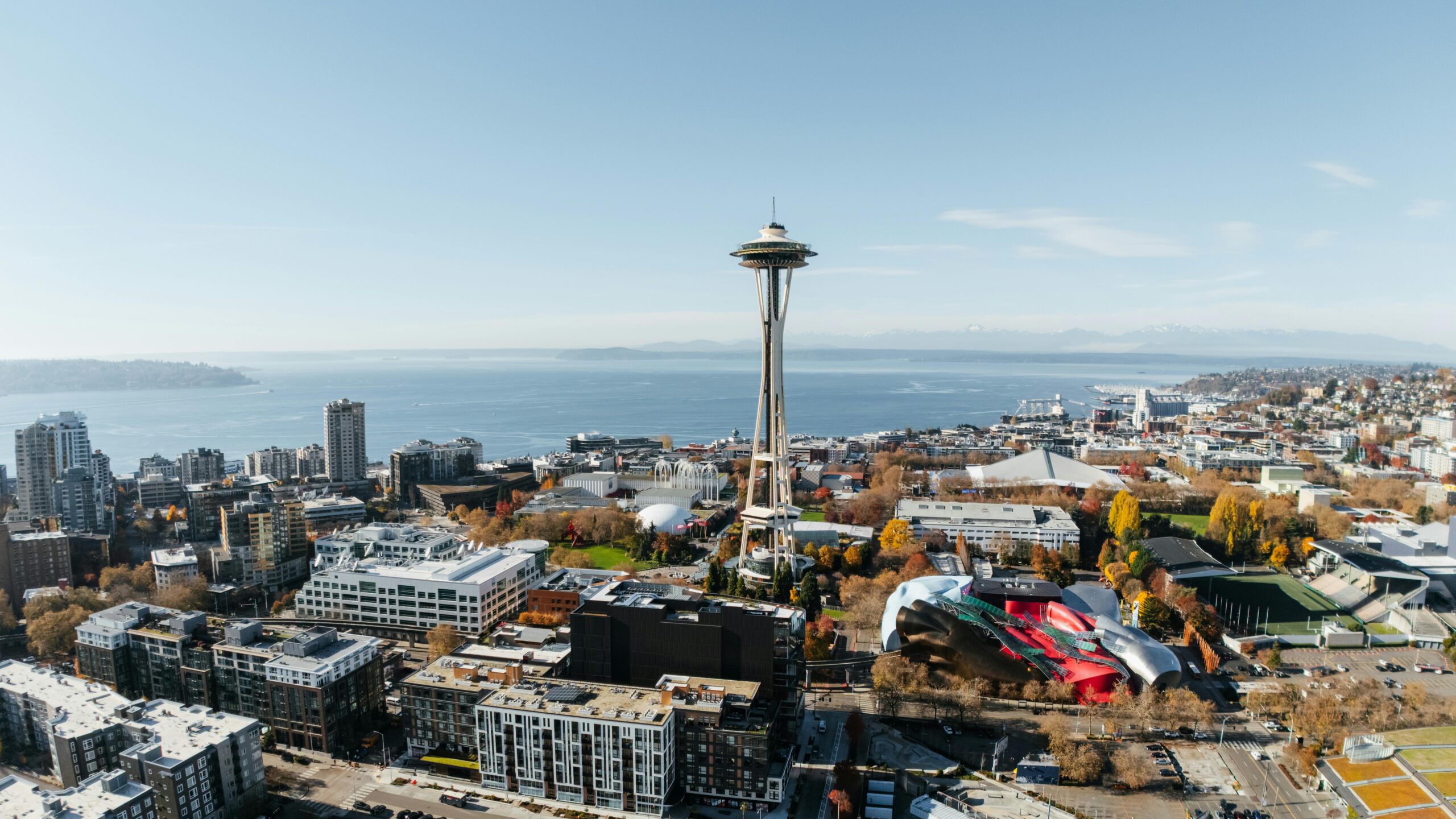 A panoramic aerial view of Seattle, Washington, showcasing the iconic Space Needle and the vibrant MoPOP (Museum of Pop Culture) building with its unique colorful facade in Seattle Center, with Elliot Bay and the Olympic Mountains visible in the background under a clear sky. British Swim School offers year-round, indoor swim lessons for the Seattle community and surrounding Queen Anne, South Lake Union, and Capitol Hill neighborhoods.