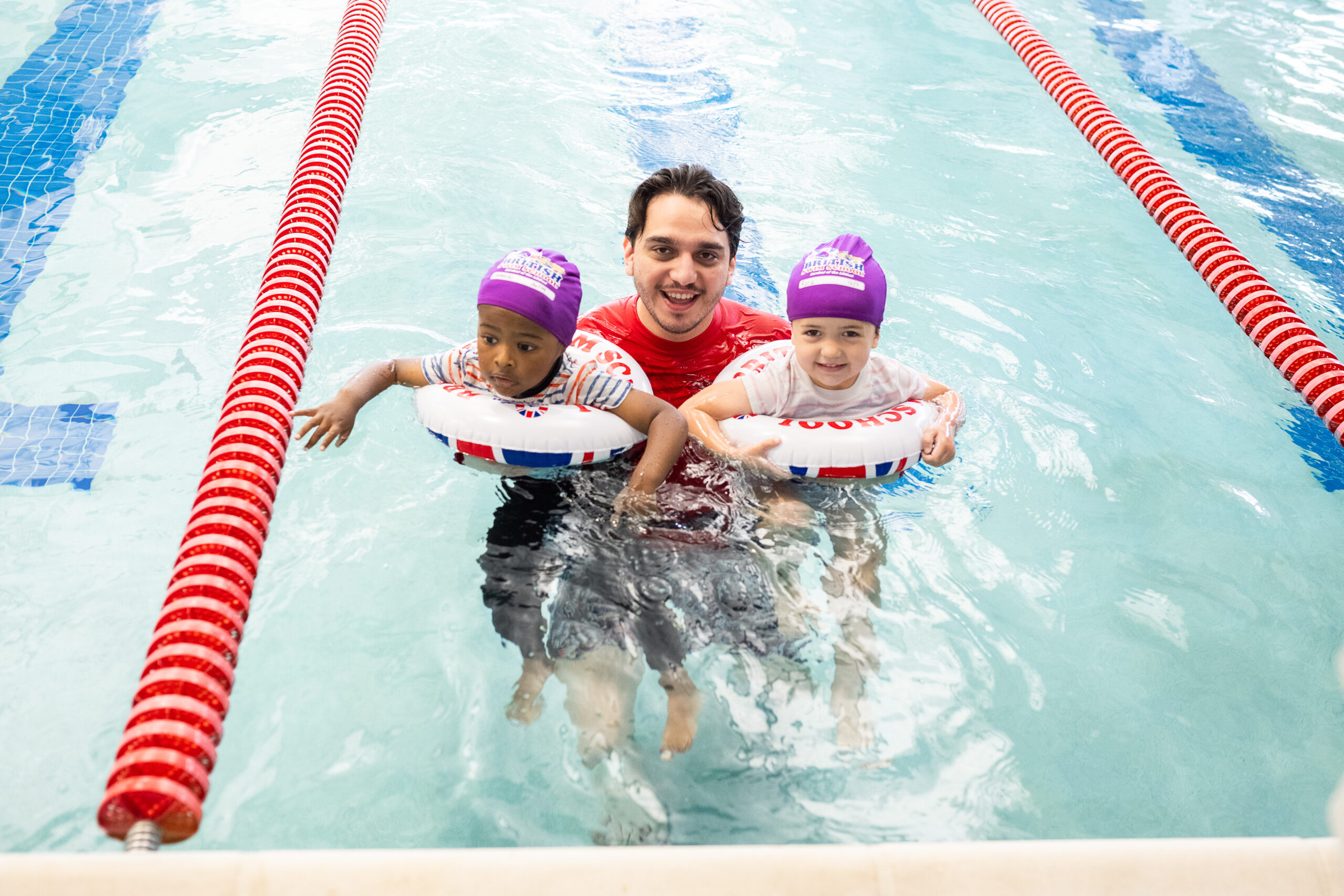 A swim instructor supports two young children wearing swim caps and flotation rings in an indoor pool during Survival Week lessons at British Swim School.