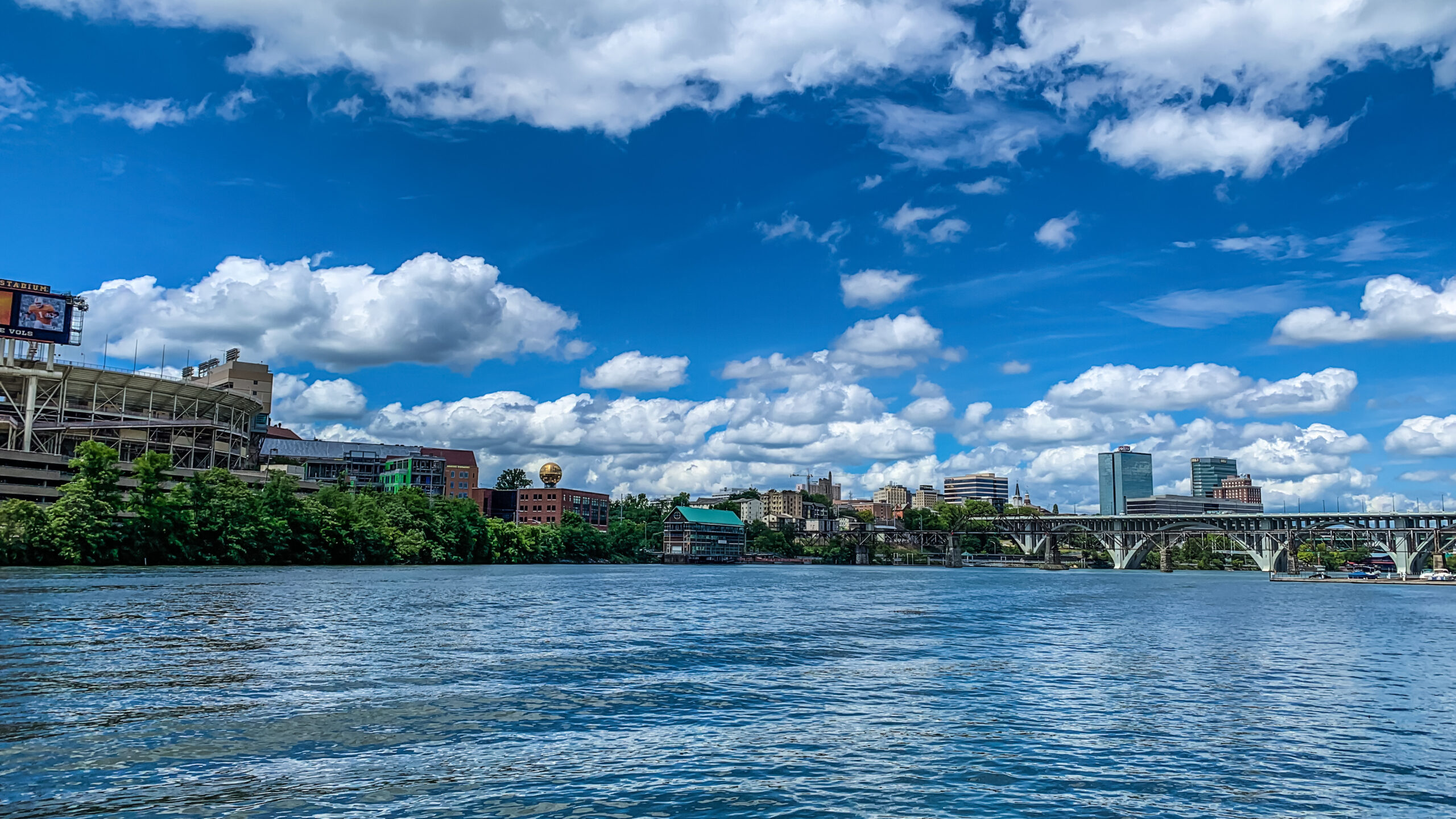 A wide-angle view across the Tennessee River toward the Knoxville, Tennessee skyline, featuring Neyland Stadium, the golden Sunsphere, and the Henley Street Bridge under a bright blue sky with fluffy white clouds.