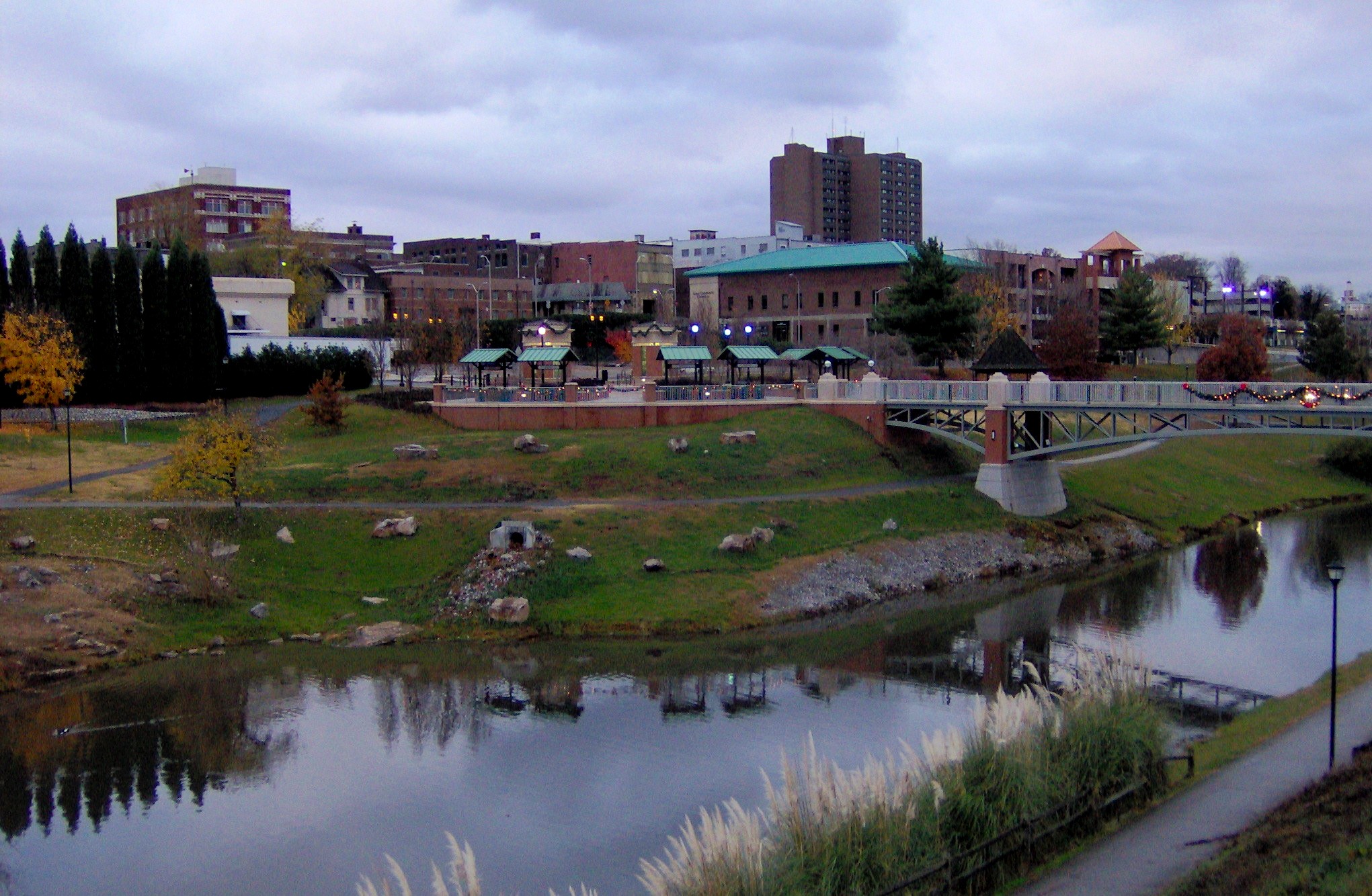 A scenic view of Bicentennial Greenbelt Park in Maryville, Tennessee, featuring a pedestrian bridge over Pistol Creek, reflection in the water, and the downtown skyline under a cloudy evening sky.