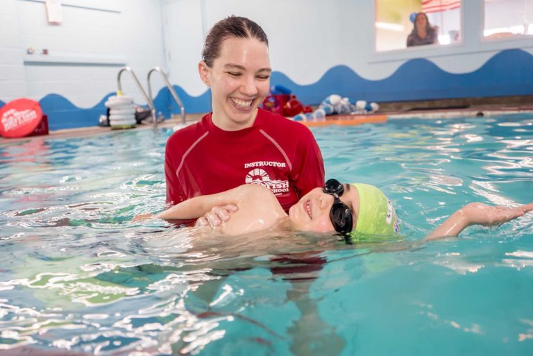 A smiling swim instructor helps a happy child float on their back during a swimming lesson.