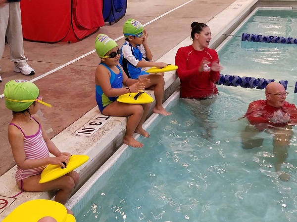 Young swimmers with kickboards sit by the pool, listening to their instructors.