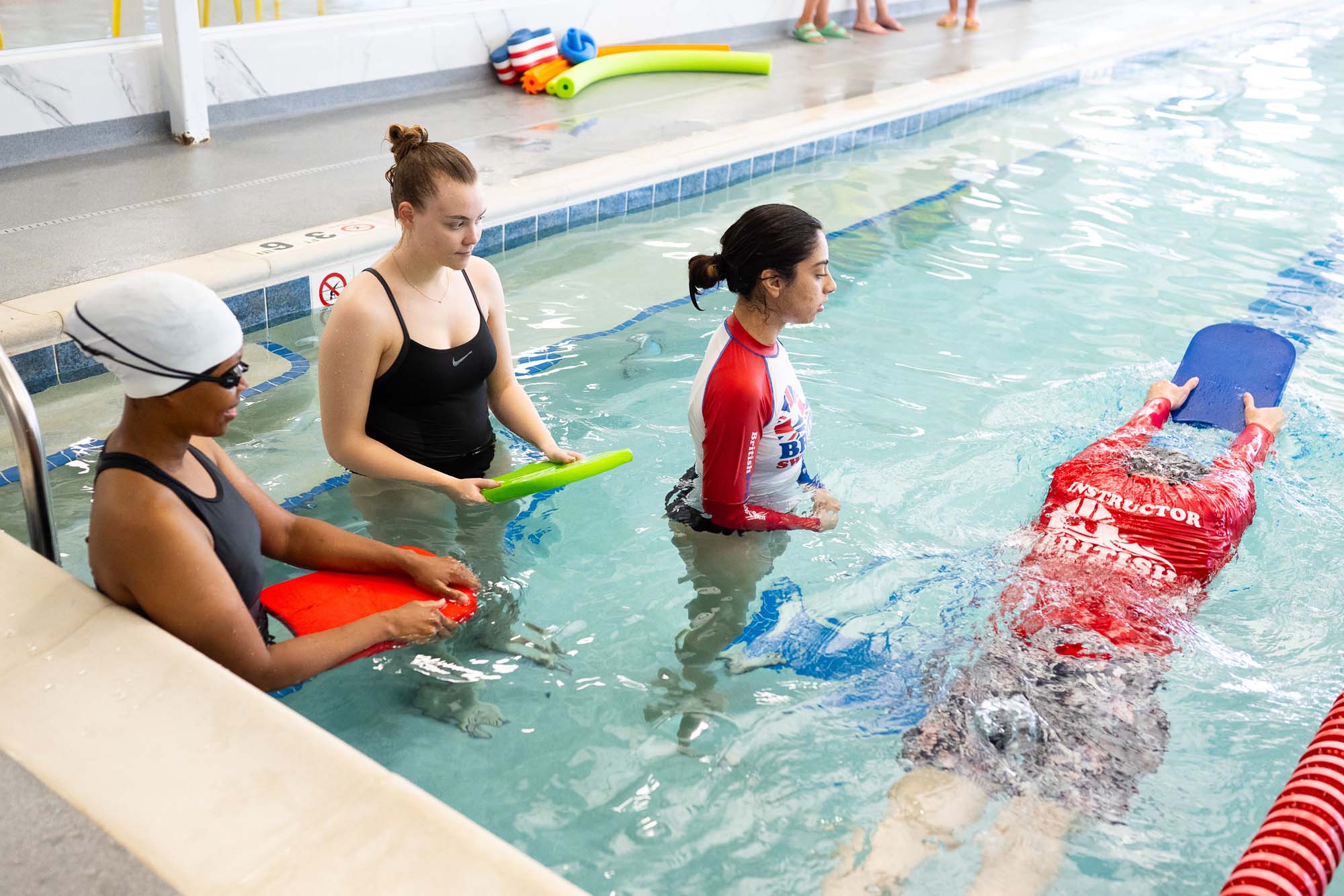 Three women are in a swimming pool, with one swimming laps and two others observing with