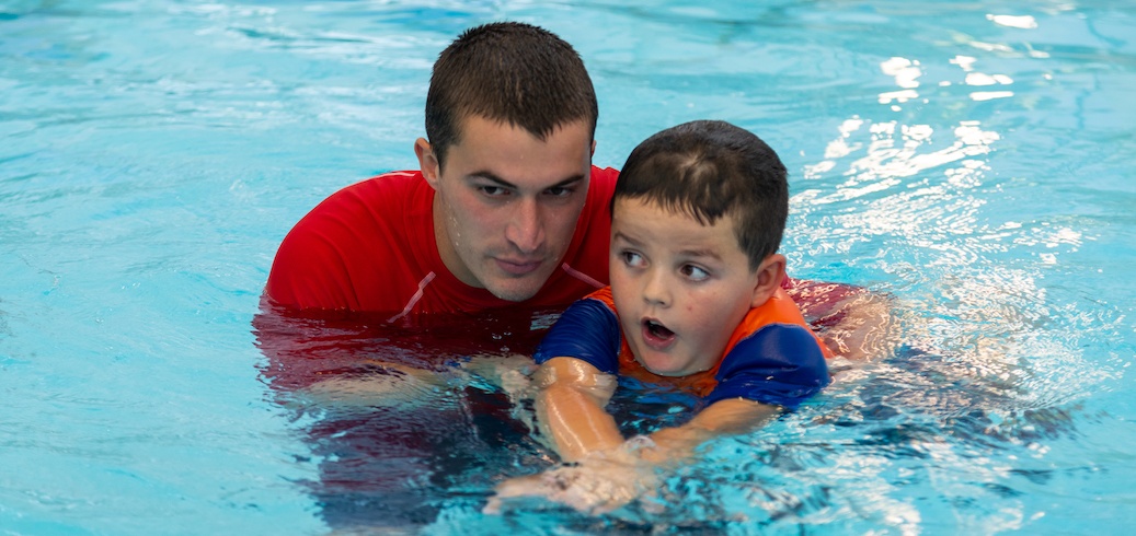A swim instructor helps a young boy learn to swim in a clear blue pool. TITLE: