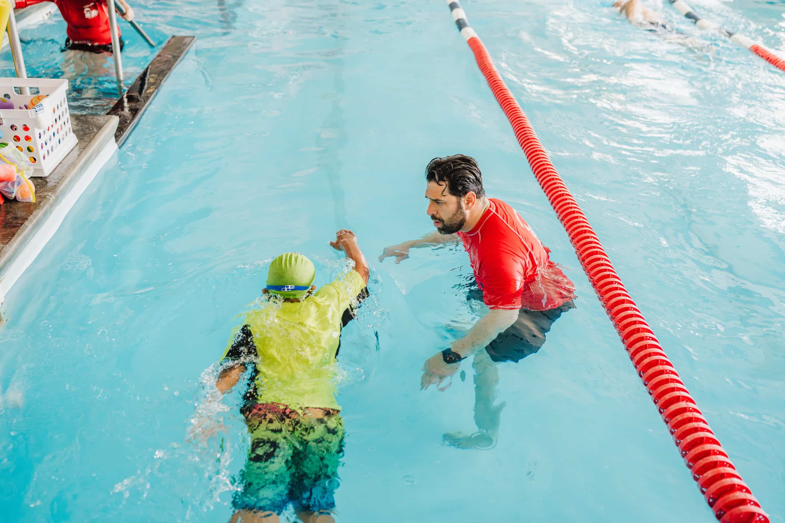 A swim instructor in a red shirt guides a child in a yellow rash guard during a swimming