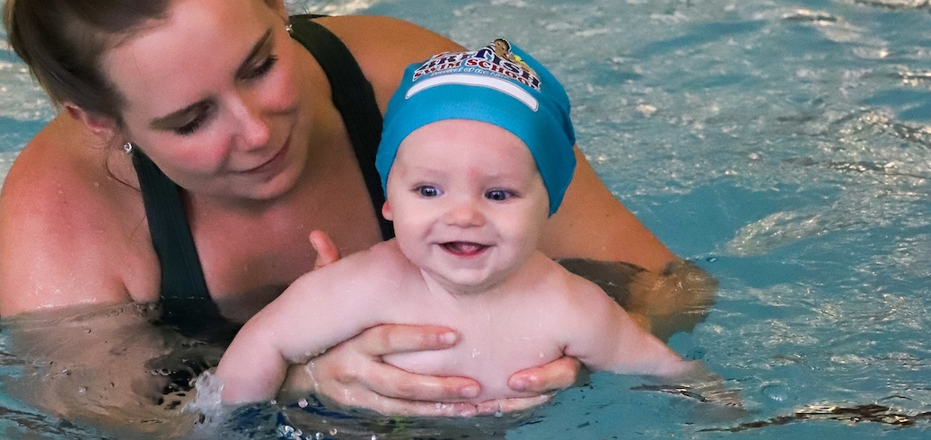 An adult gently holds a smiling baby wearing a blue swim cap in a swimming pool. TITLE