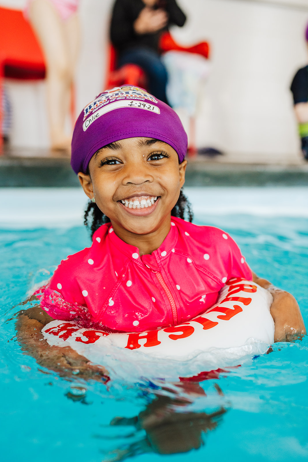 A smiling young girl in a pink swimsuit and purple cap floats happily in a swimming pool.