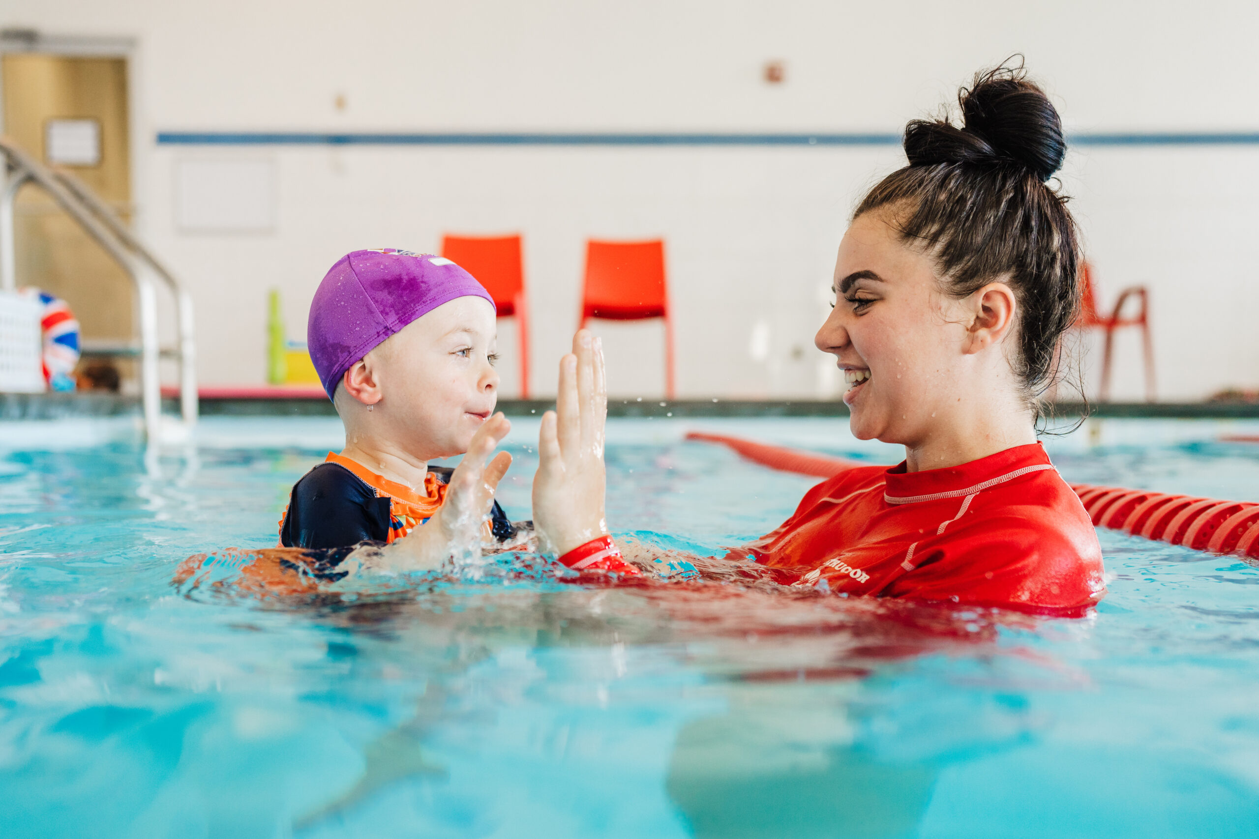A happy child in a purple cap high-fives their smiling swim instructor in a pool.