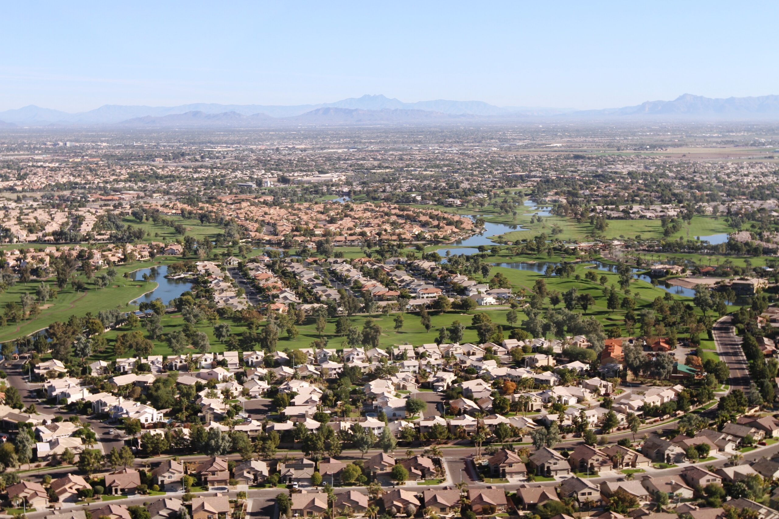 An expansive aerial photograph of a sprawling residential community in Chandler, Arizona, featuring neat rows of tiled-roof houses built around lush green golf course fairways and a long, sinuous lake system under a clear sky with distant mountains. British Swim School offers year-round, indoor swim lessons for the Maricopa County community and surrounding Chandler, Gilbert, Tempe, and Mesa neighborhoods.