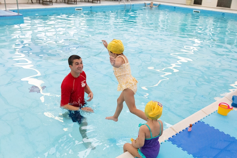 A swim instructor guides a child jumping into an indoor pool as another child watches.
