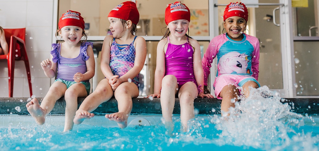 Four happy girls in red swim caps sit by a pool, splashing water with their feet.