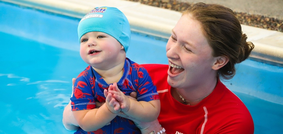 A smiling baby in a blue swim cap is held by a laughing instructor in a pool