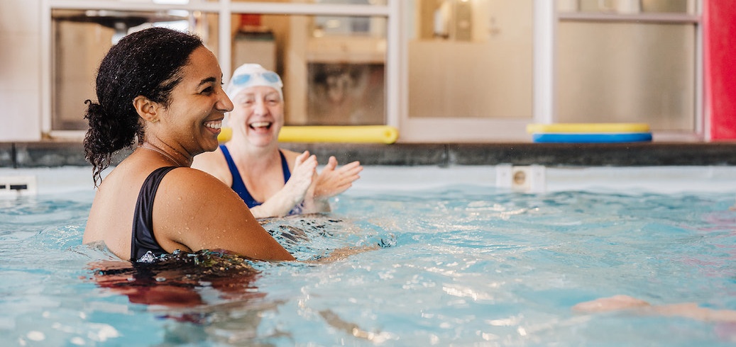 Two diverse women happily swim and clap during an indoor pool class.