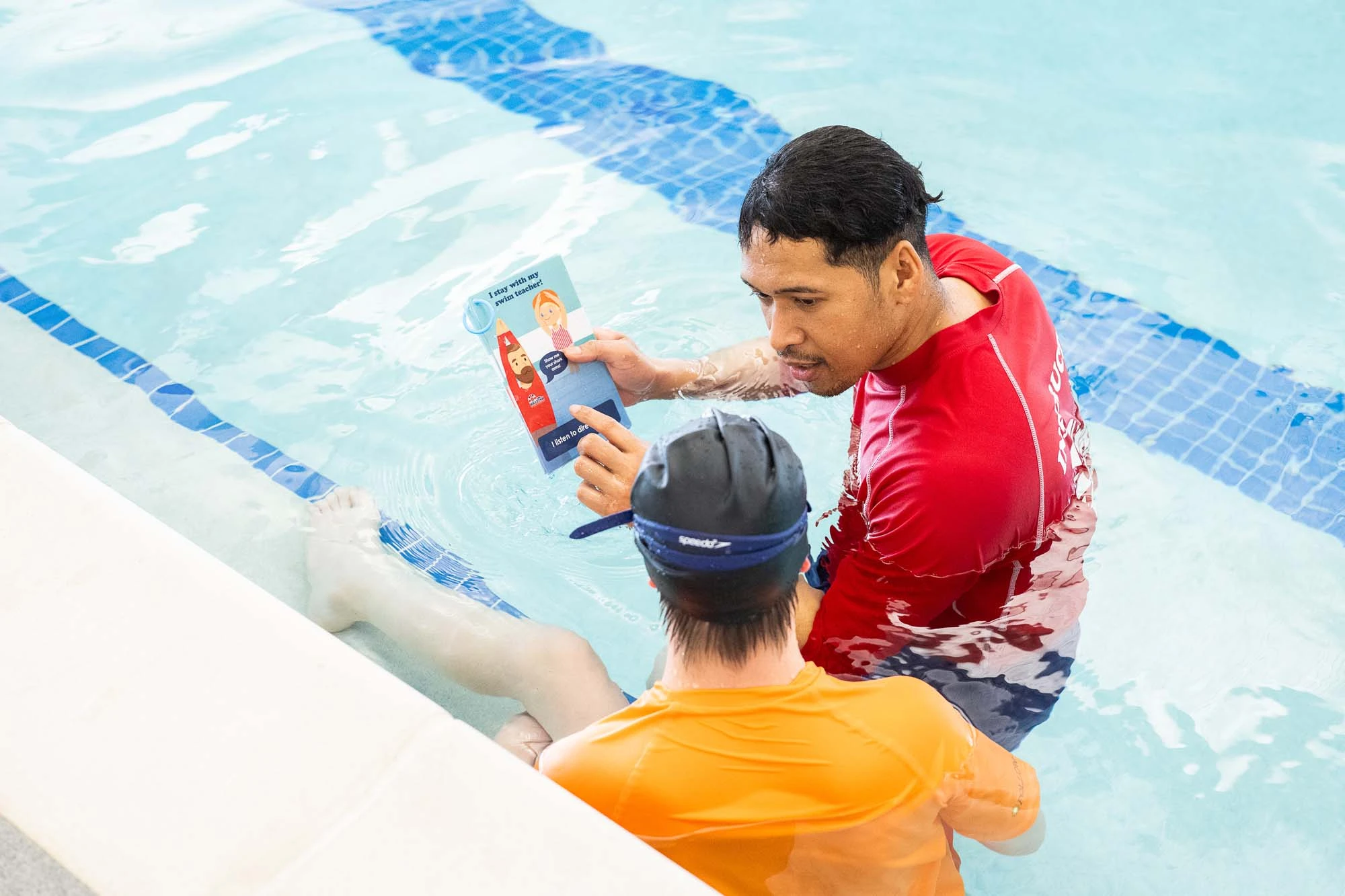 A swim instructor in red shows a booklet to a student in orange and a swim cap in