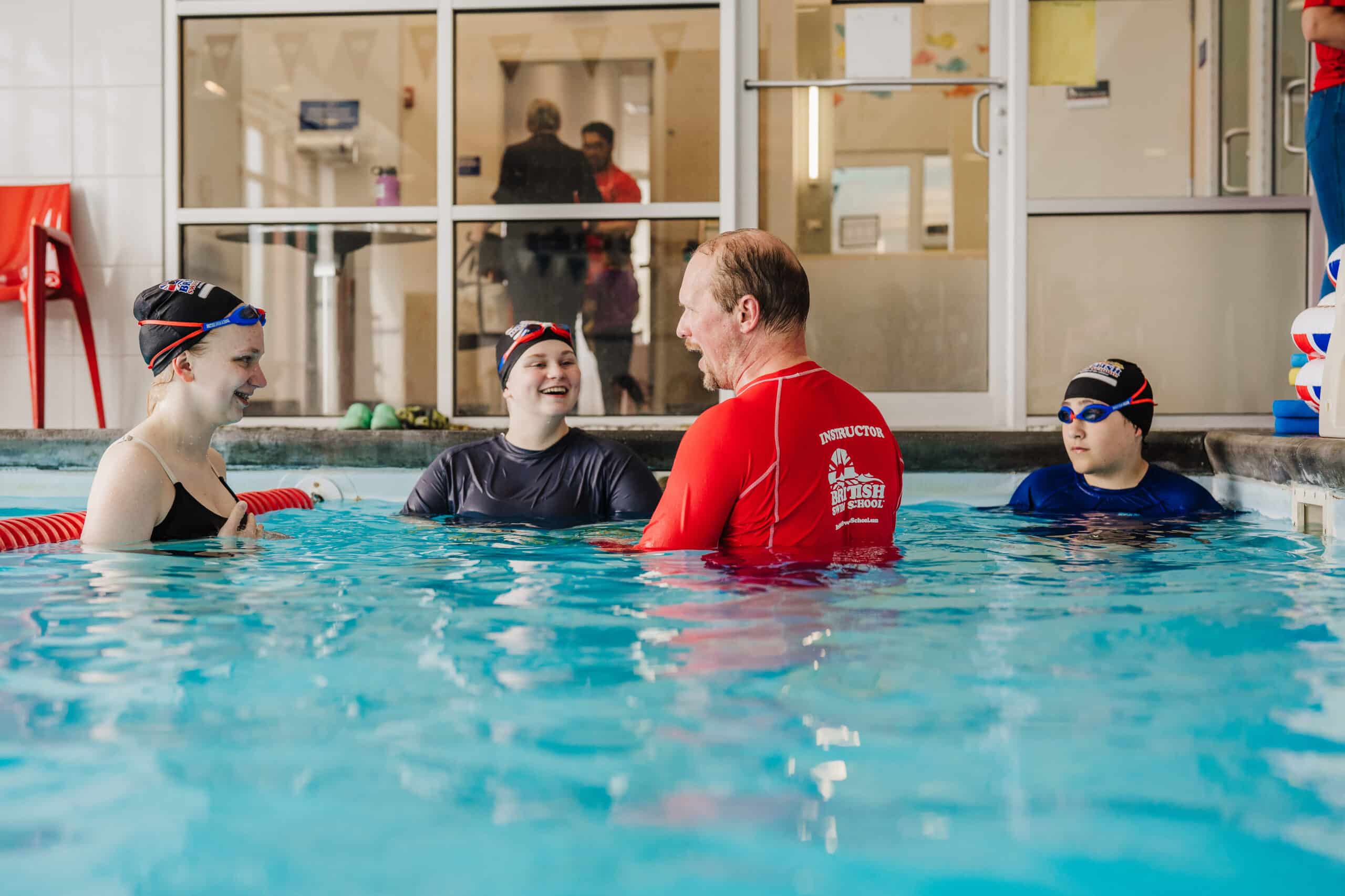 An adult swim instructor teaches three smiling children in a clear indoor pool.