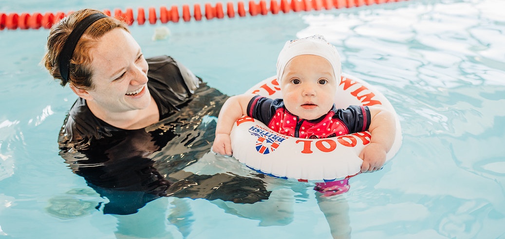 A smiling woman watches a baby in a swim float during a swimming lesson.