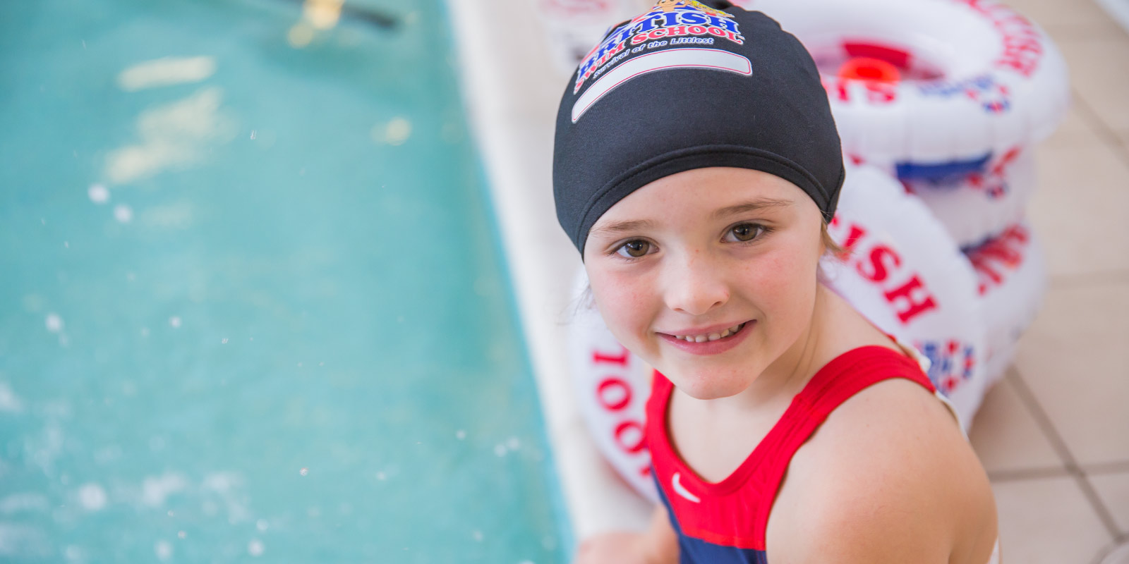 A smiling young girl in a black swim cap and red swimsuit sits by a swimming pool
