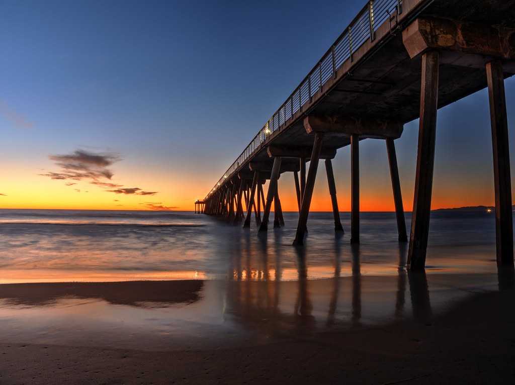 A low-angle view from the sand underneath the Hermosa Beach Pier in California at sunset, showing the long wooden structure and pilings stretching into the ocean against a glowing orange and deep blue twilight sky.