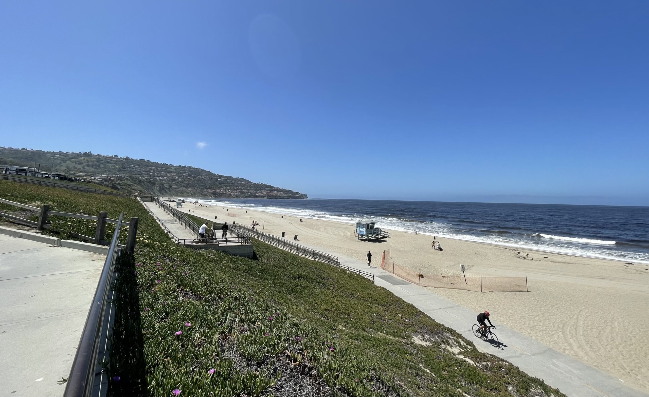 A sunny, wide-angle view of Torrance Beach in California, featuring a paved bike path with a cyclist, a lifeguard stand on the sand, and the Palos Verdes Peninsula in the background under a clear blue sky.