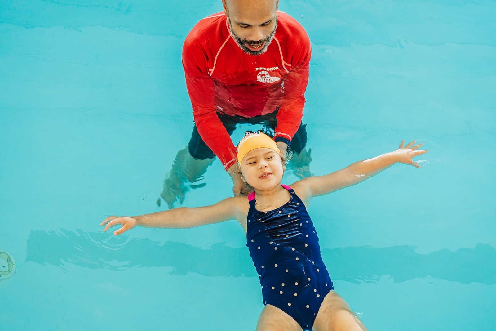 A swimming instructor supports a young girl floating on her back in a bright blue pool.