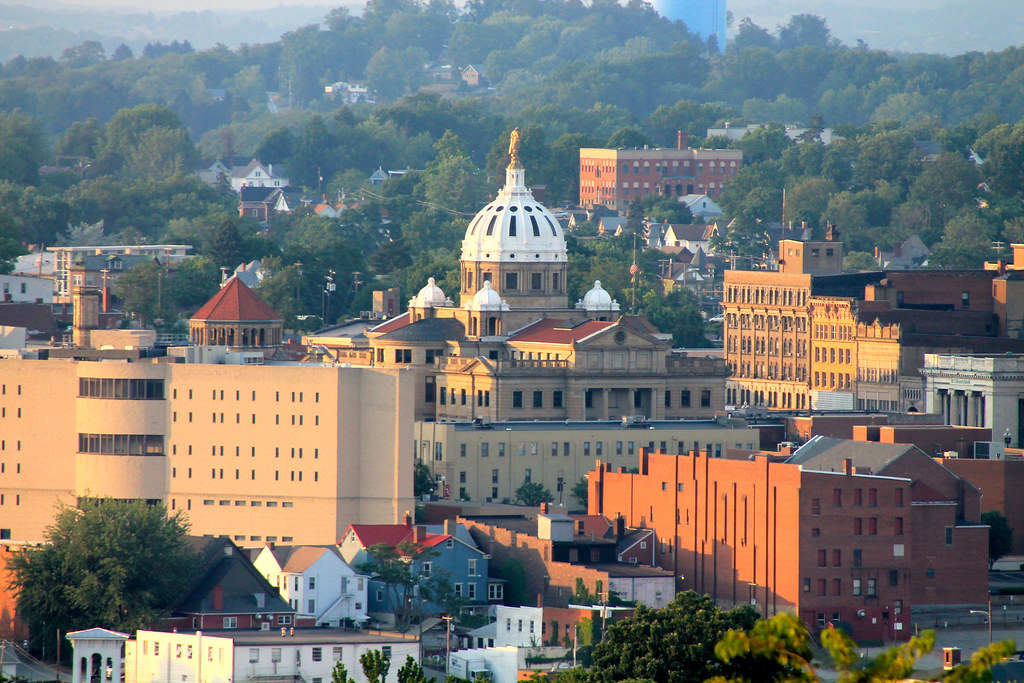 An elevated view of downtown Washington, Pennsylvania, featuring the prominent white dome and golden statue of the Washington County Courthouse, surrounded by historic brick buildings and rolling green hills at sunset.