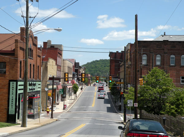 A sunny, street-level view of Pike Street in downtown Canonsburg, Pennsylvania, featuring historic brick storefronts, Humble Carpet shop, traffic lights, and a lush green hill in the distance under a blue sky.
