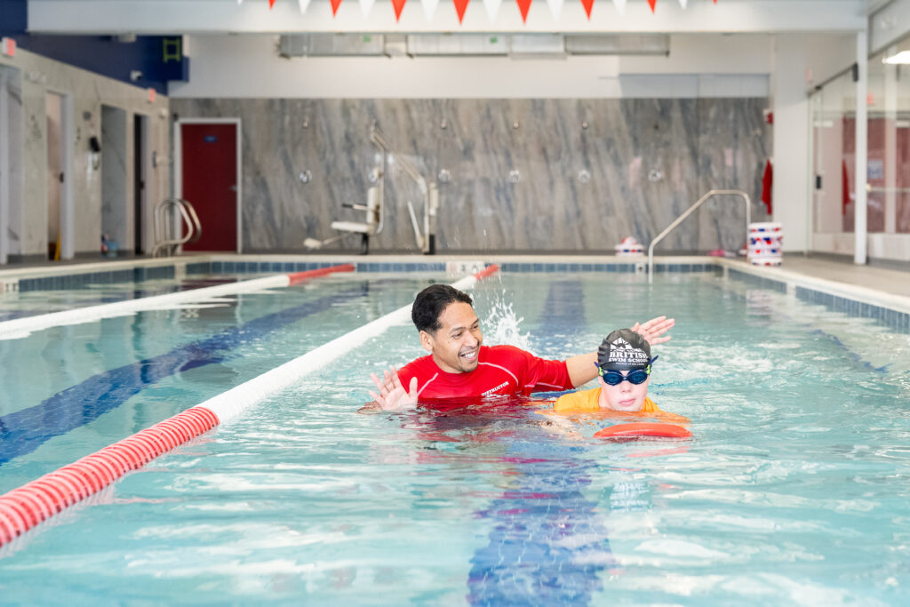 Instructor supporting a young swimmer with a kickboard during a British Swim School adaptive aquatics lesson.