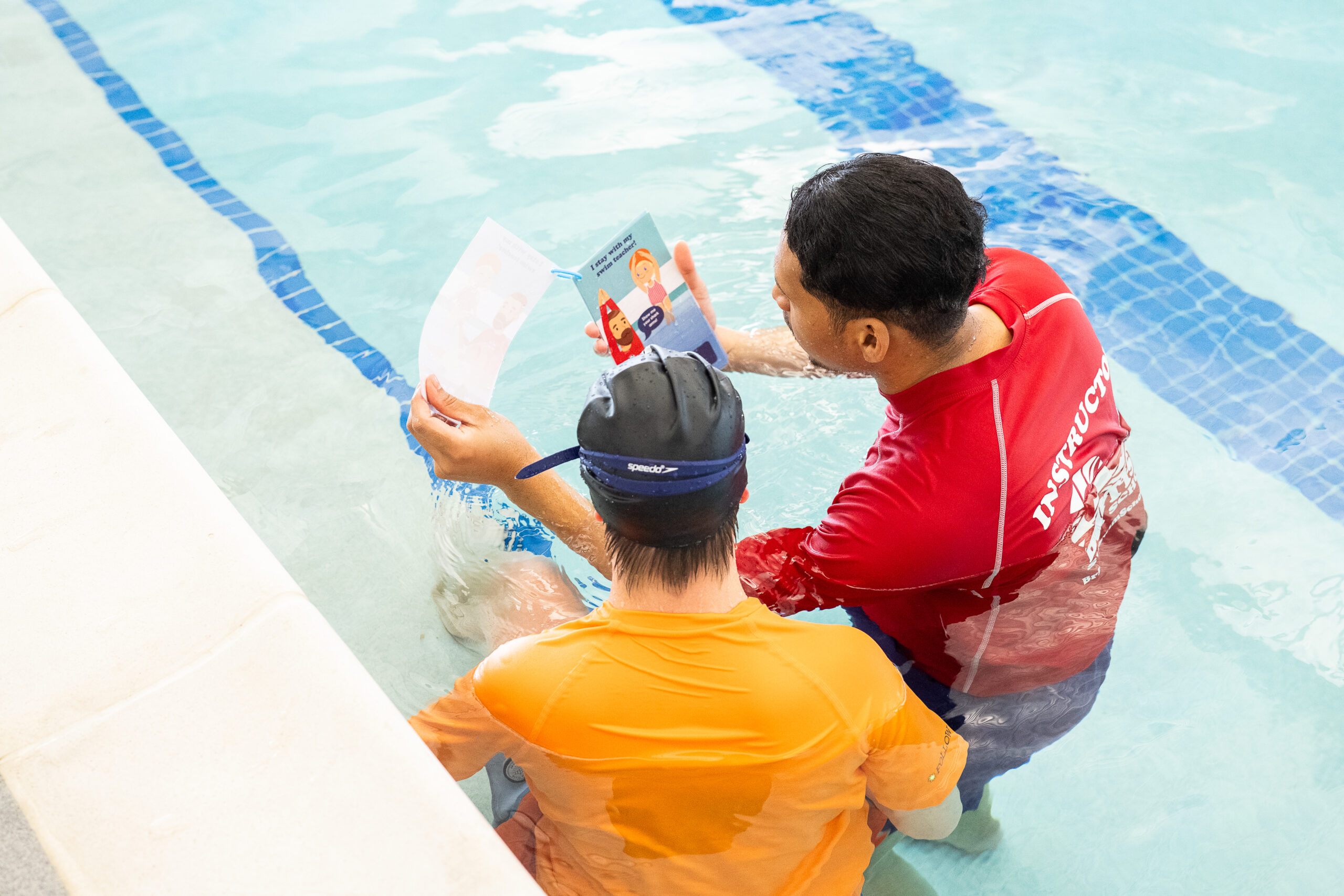 Swim instructor showing a young student illustrated safety cards during an adaptive aquatics swim lesson at British Swim School.