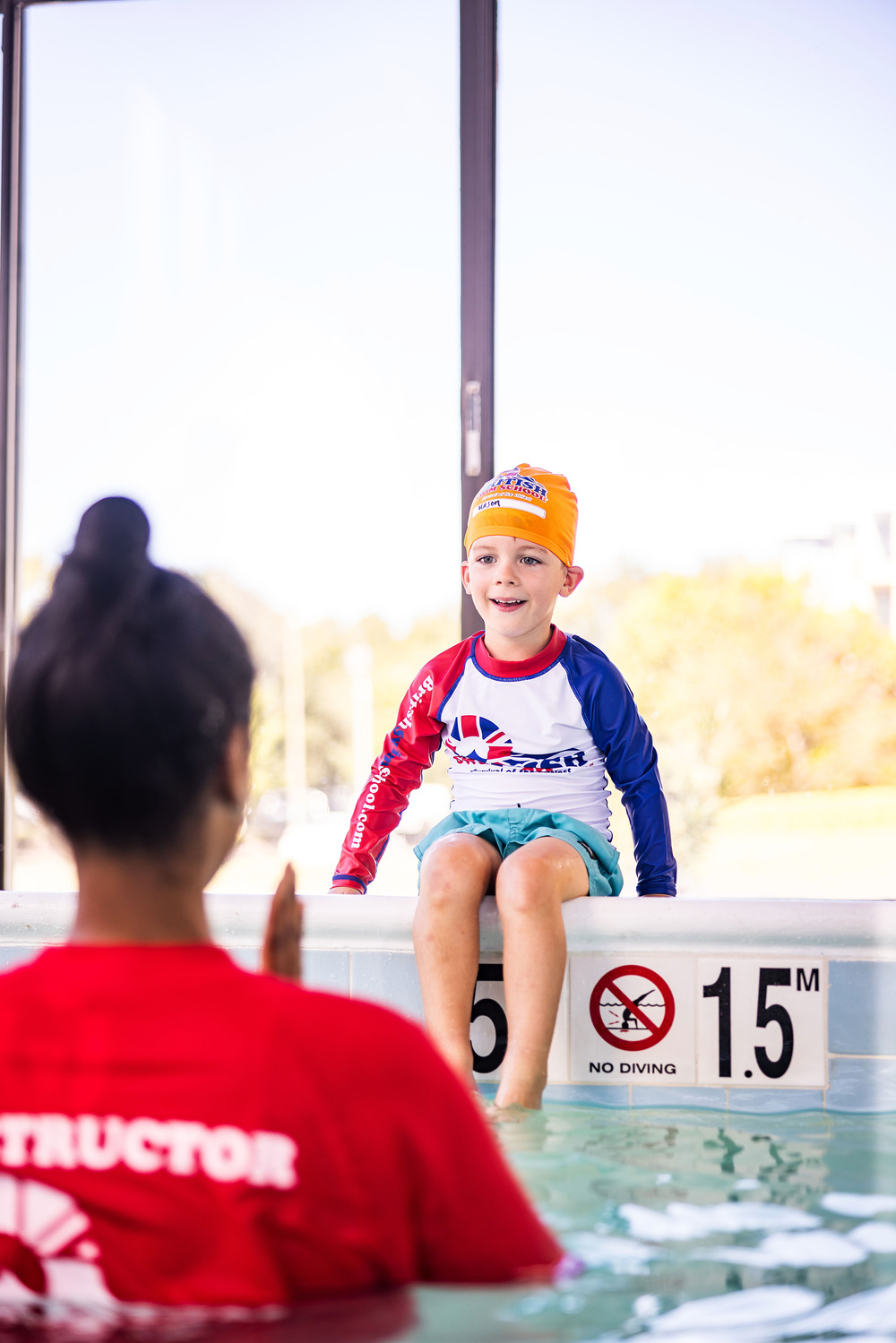 A young boy wearing an orange swim cap sits on the pool edge during a British Swim School swim lesson with his instructor.