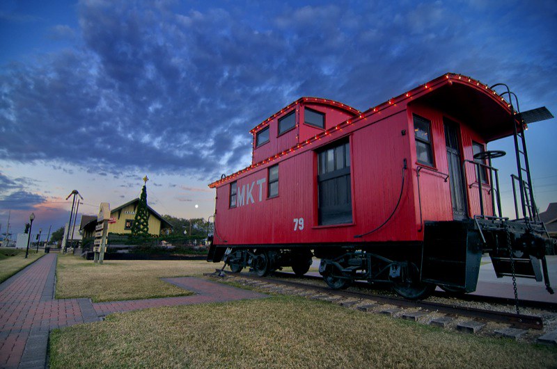 A low-angle view of a bright red MKT caboose decorated with Christmas lights at the historic Katy Depot in Katy, Texas, under a dramatic twilight sky with the moon visible.