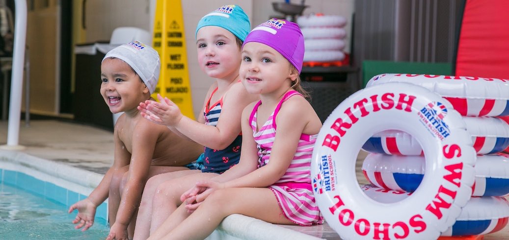 Smiling children in swim caps sit at the pool's edge with British Swim School floats