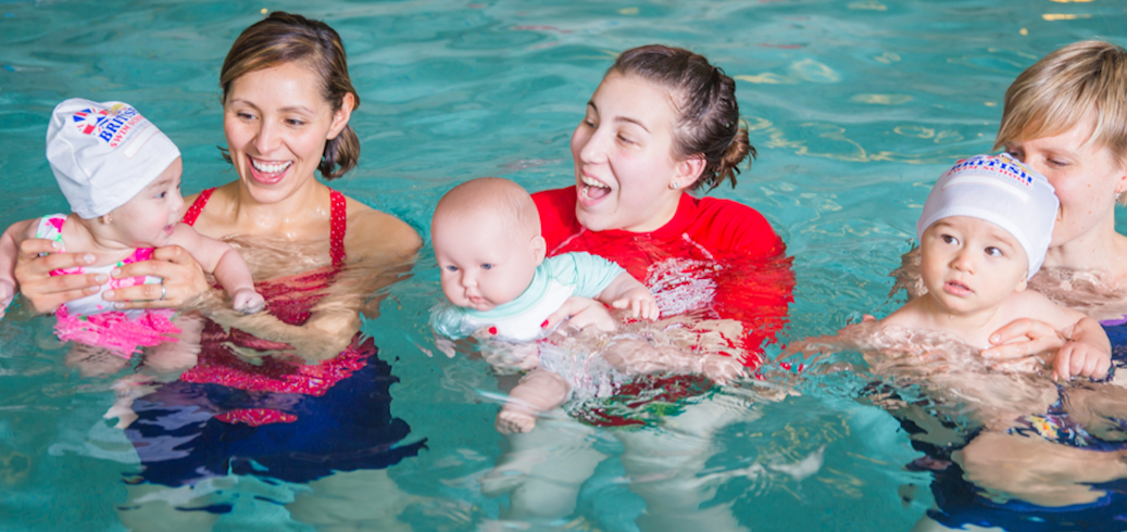 Parents and an instructor happily guide three babies during a swim class. TITLE: