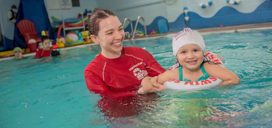 A smiling swim instructor helps a happy young girl wearing a swim cap and floatie in a pool. TITLE