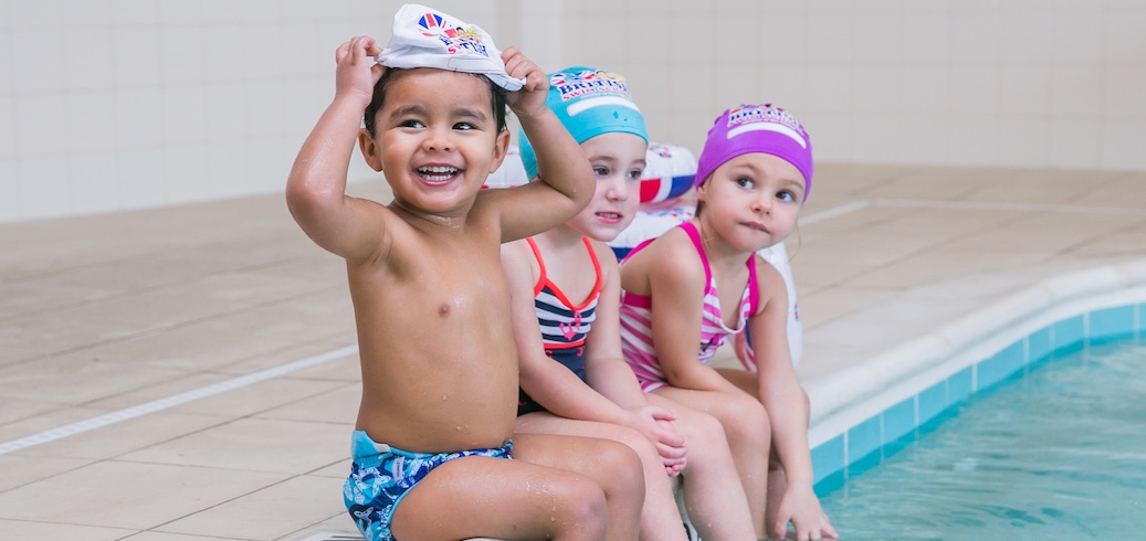Three smiling young children in swimsuits and caps sit at the edge of a swimming