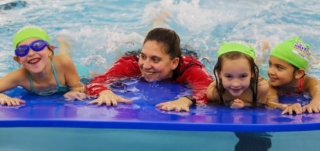 A smiling swim instructor helps three happy girls on a blue floating mat during a swim lesson.