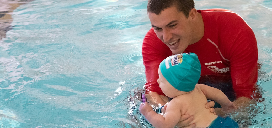 A smiling instructor teaches a baby in a teal swim cap during a swimming lesson. TITLE