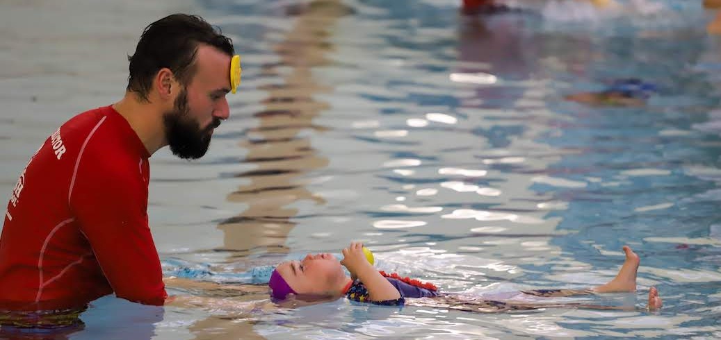 A swim instructor supervises a child learning to back float in a swimming pool. TITLE: