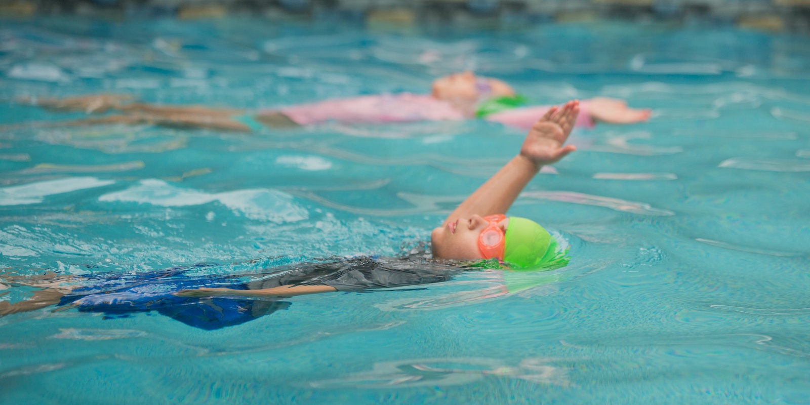 A child wearing a green swim cap and orange goggles floats on their back in a pool with