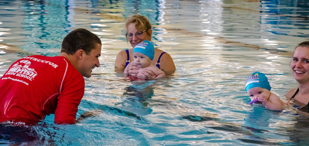 A swim instructor smiles at two women and two babies in a pool during a swim