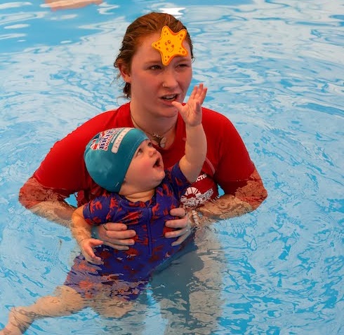 A woman with a starfish on her forehead holds a baby in a blue swimming pool. TITLE