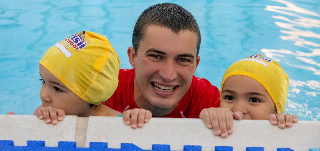 A smiling swim instructor and two children in yellow caps peek over the pool edge. TITLE