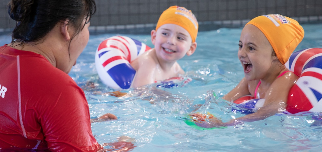 Happy children in orange swim caps and Union Jack floaties splash with an instructor in a