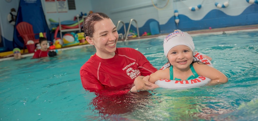 A smiling swim instructor helps a happy young girl in a swim ring during a lesson. TITLE